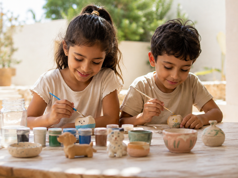 Two children painting their finished clay pieces at a sunlit table