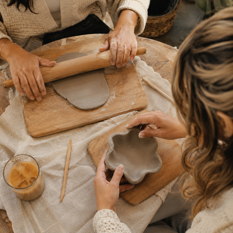 Overhead view of two people rolling clay and shaping a flower-shaped dish on a wooden board