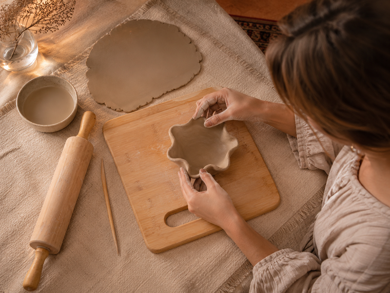 Overhead view of hands shaping a wavy clay bowl on a wooden board